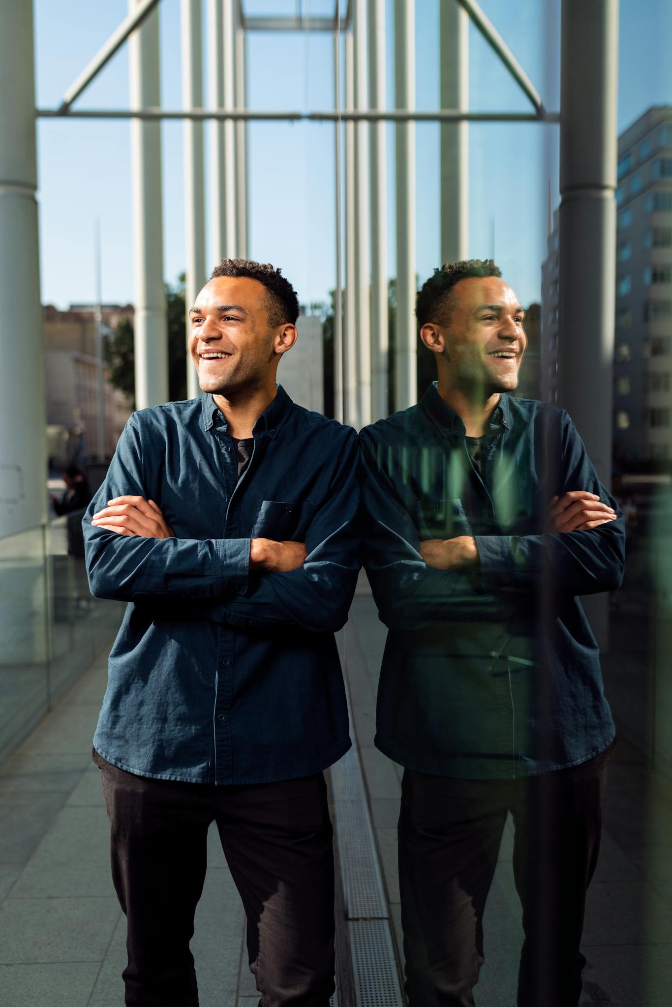 A confident male professional with arms crossed, reflecting against a glass wall, enjoying a sunny day.